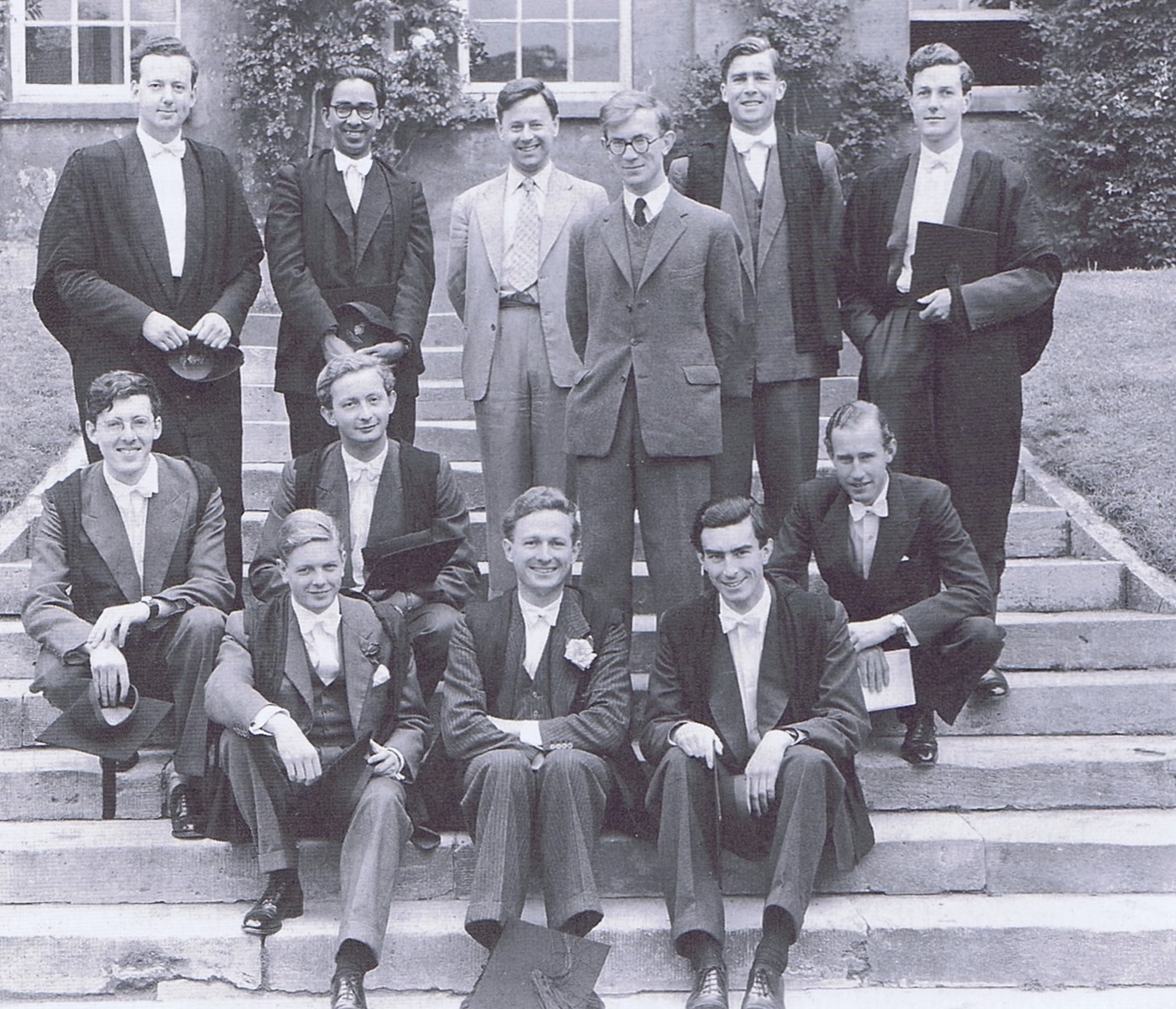 Black and white photograph of young men standing and sitting on the steps of Worcester quad. They are wearing sub-fusc and academic gowns.