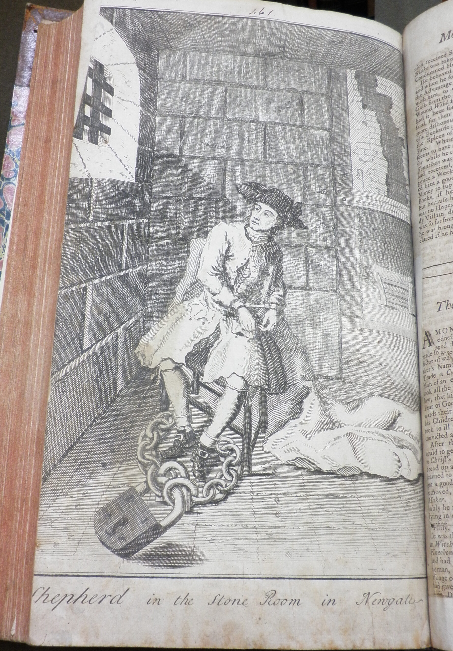 A man in 18th century dress sits in a stone prison cell. An exaggerated large chain and lock are around his ankles.